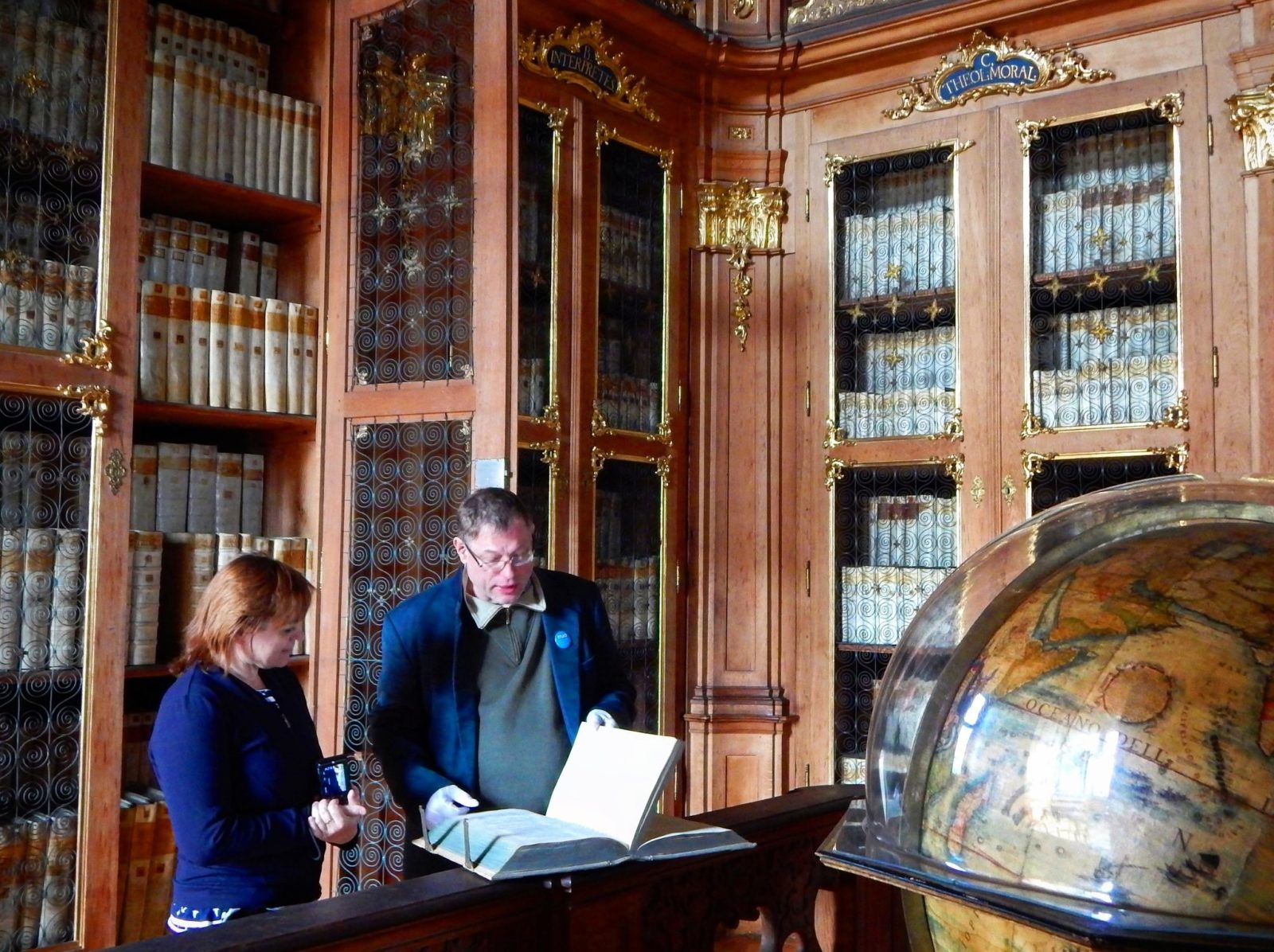 Female and male historians stand reviewing a book in a library. Globe is in view.